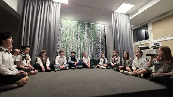 Grupa uczniów śpiewa siedząc na scenie w półokręgu